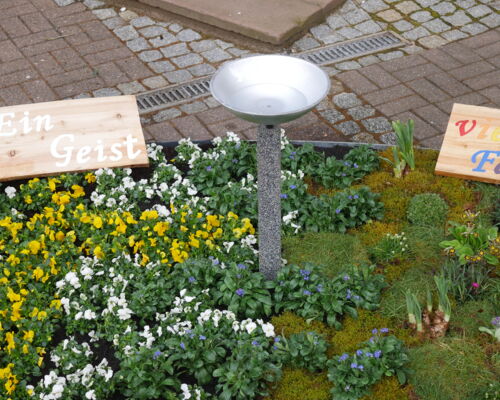 Das Zentrum des Blumenbeetes. Kreuz und Wiese sind durch wellenförmig gepflanzte blaue Blumen verbunden. In der Mitte steht ein Wasserbecken auf einer Stele. Eine Holztafel trägt die Aufschrift: Taufe verbindet.