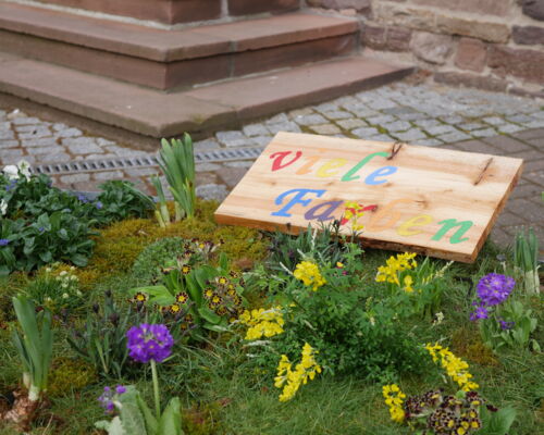 Detailansicht des Blumenbeetes, es zeigt verschiedene Blumen. Auf einer Holztafel steht in bunten Buchstaben: viele Farben.